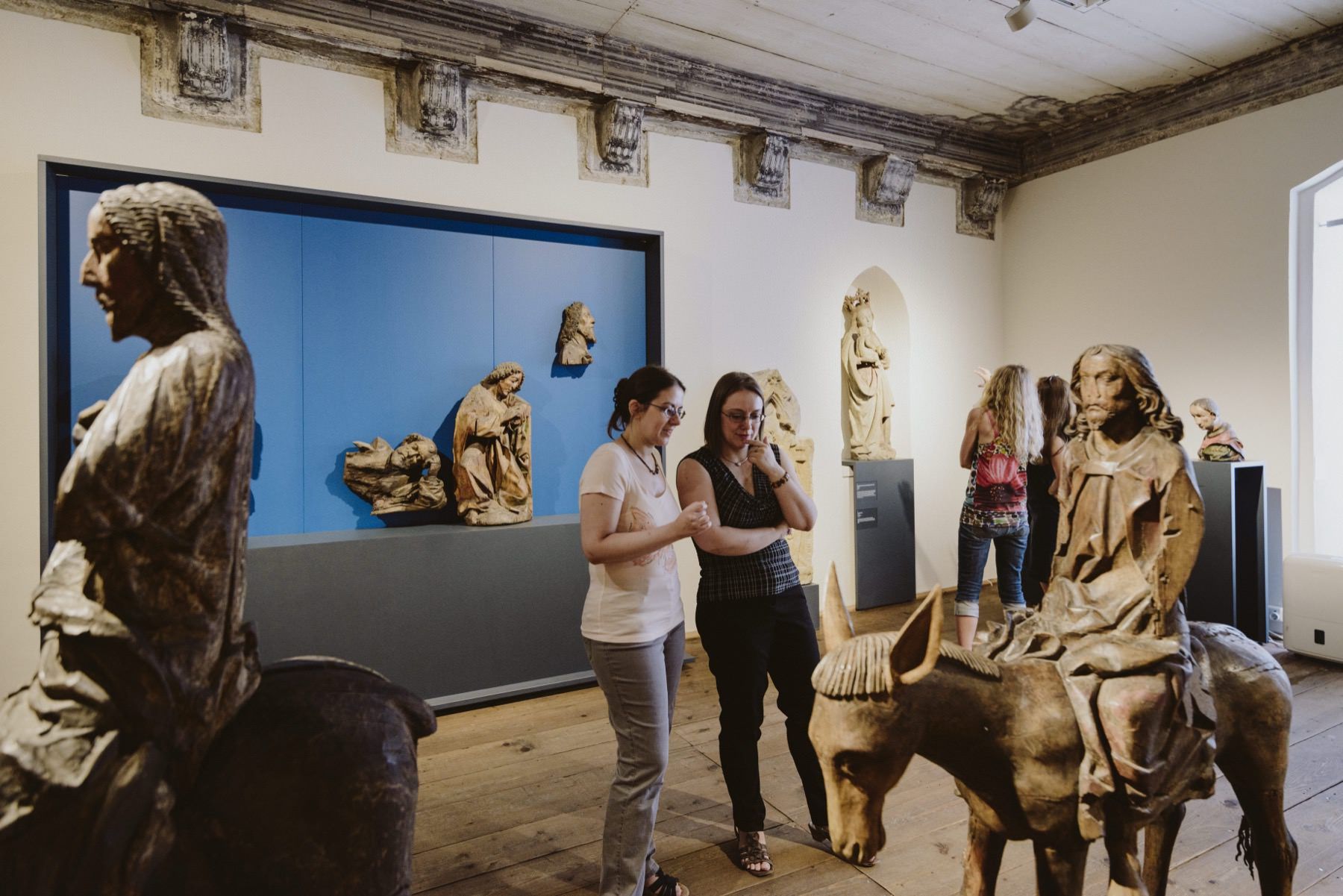 Ausstellungsraum mit historischen Figuren (eine Person reitet auf einem Pferd, eine reitet auf einem Esel). In der Mitte sthen 2 Personen und betrachten die Statuen.